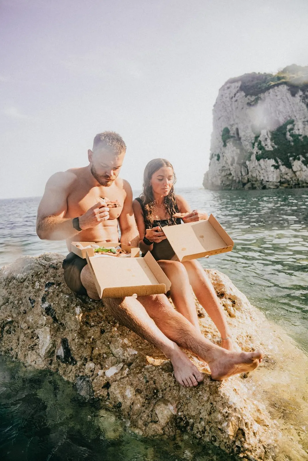 Two people eating takeaway on a rock with chalk stacks behind them.